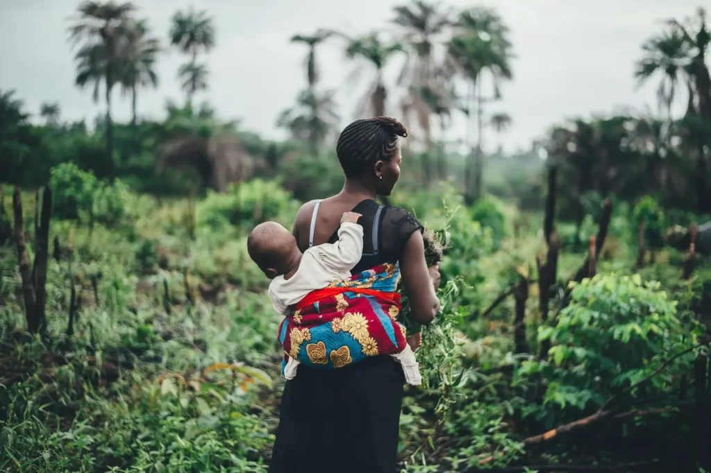 A woman in a black dress with a baby strapped to her back looks at a field. Her back is to the camera and both her and her baby’s face is not visible.