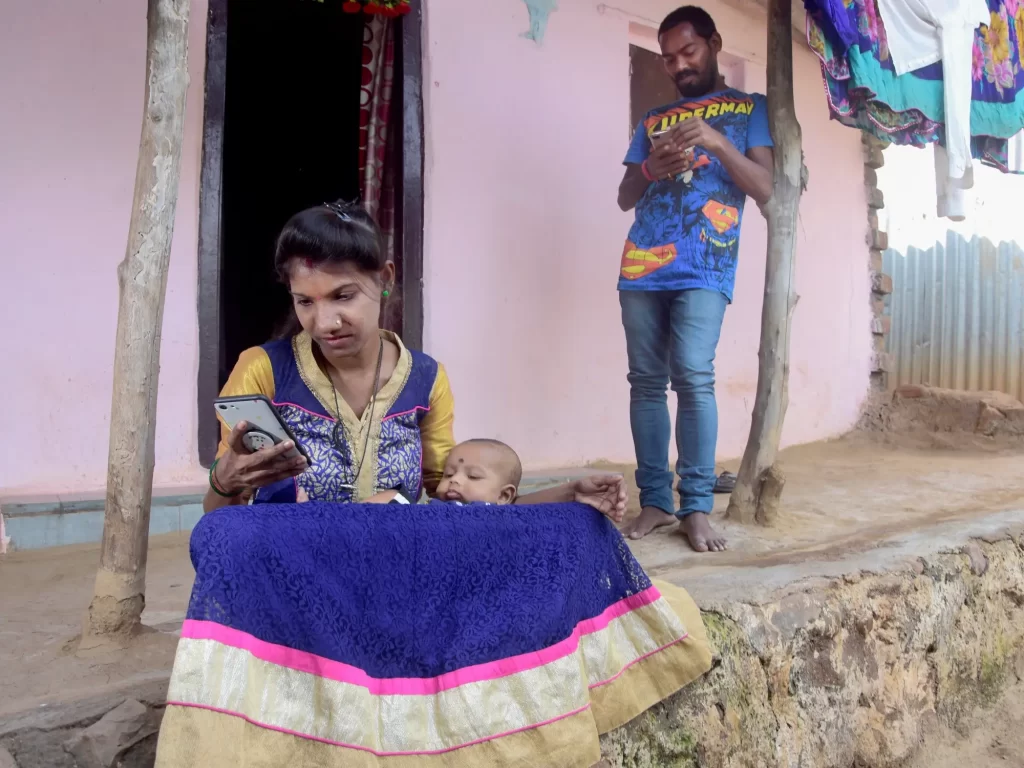 A woman in a blue dress scrolls through a phone as she sits outside a house with a baby on her lap. Behind her is a man in a blue shirt and jeans, standing and looking at his phone. The house’s facade is pink and there are clothes drying in the background.